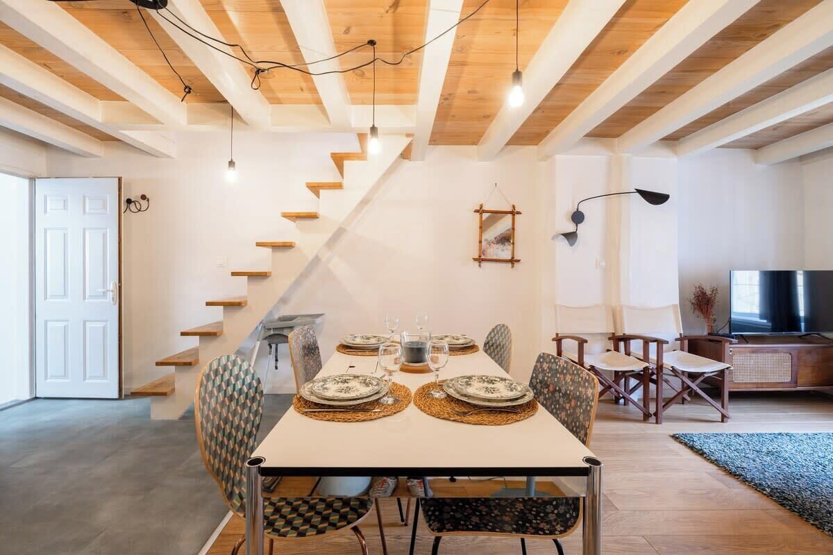 A dining area with a table set for four, wooden beams on the ceiling, and a staircase leading to another room.