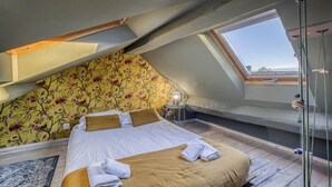 Un dormitorio con techo inclinado, una cama con ropa blanca, un papel tapiz floral y un tragaluz.