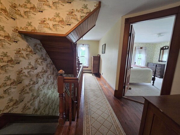 A hallway with patterned wallpaper, a wooden staircase, and a view into a bedroom with a bed and dresser.