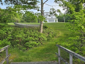 A wooden bridge leading to a grassy area with a boat and a white house in the background.