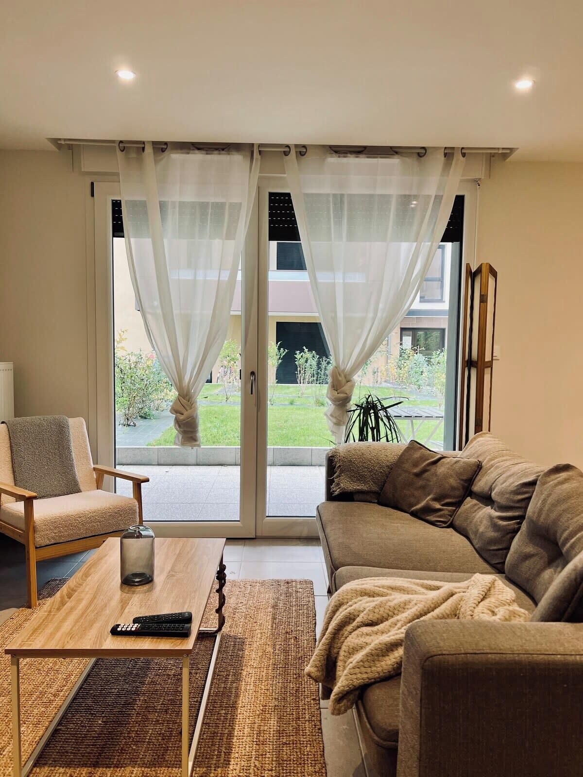 A living room with a sofa, armchair, and a coffee table. There is a view of a garden through a glass door.