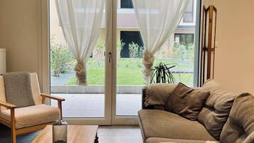 A living room with a sofa, armchair, and a coffee table. There is a view of a garden through a glass door.