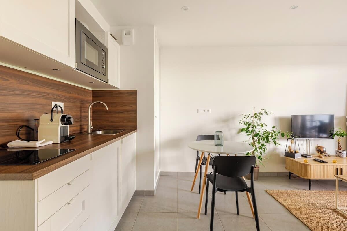 Een moderne keuken met een magnetron, waterkoker en broodrooster. Er is een eethoek met een tafel en stoelen. Een televisie is aan de muur bevestigd.