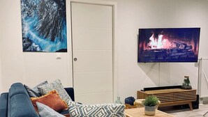 A living room with a blue sofa, a wooden coffee table, a TV displaying a fireplace, and a door. - Cozy T2 (1-bedroom) with vineyard view (Rosheim)