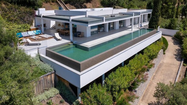 Ein modernes Haus mit großem Schwimmbad, einer überdachten Terrasse und Blick auf die umliegende Natur.