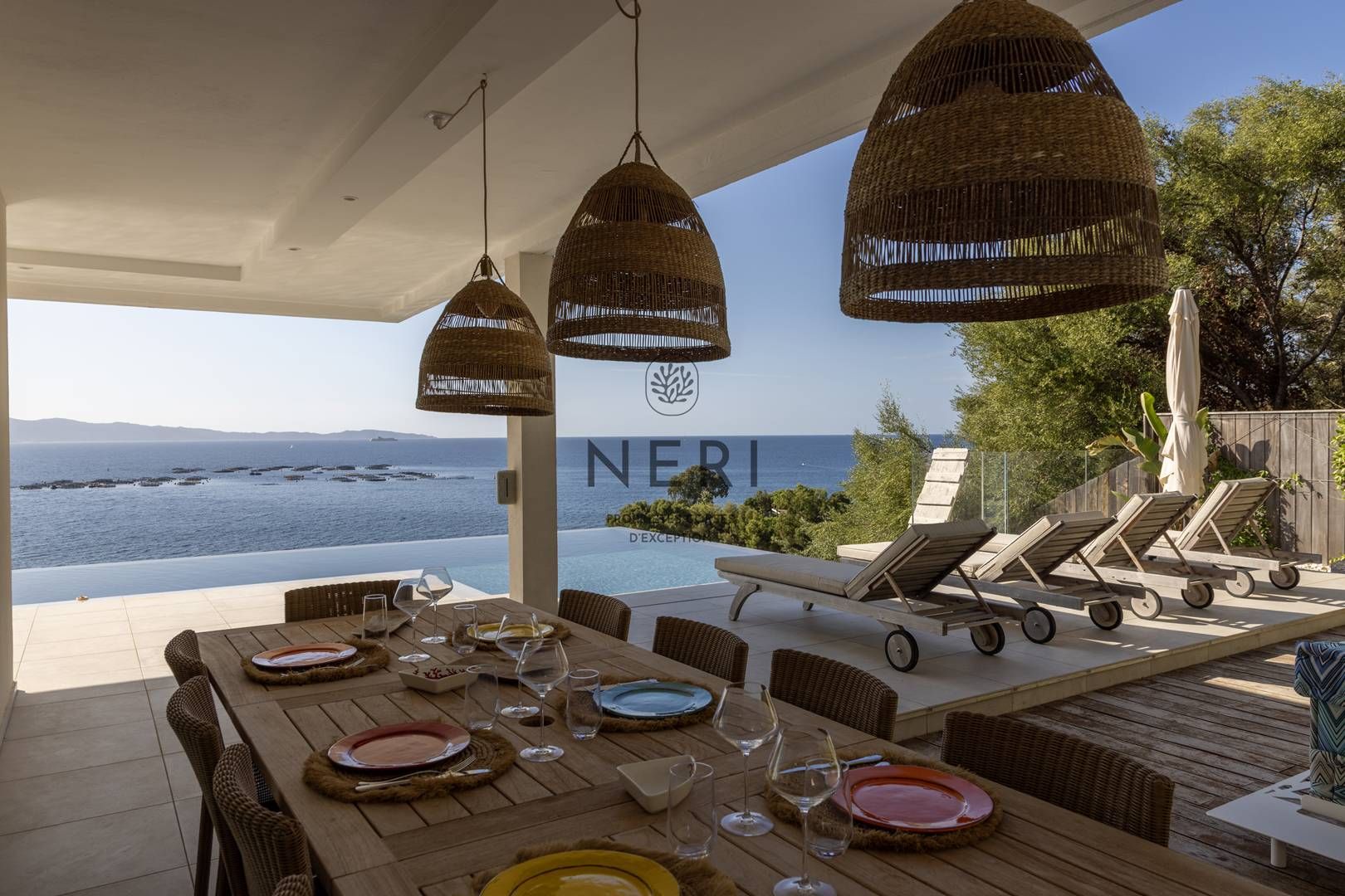 An outdoor dining area with a view of the sea, wicker pendant lights, and lounge chairs.
