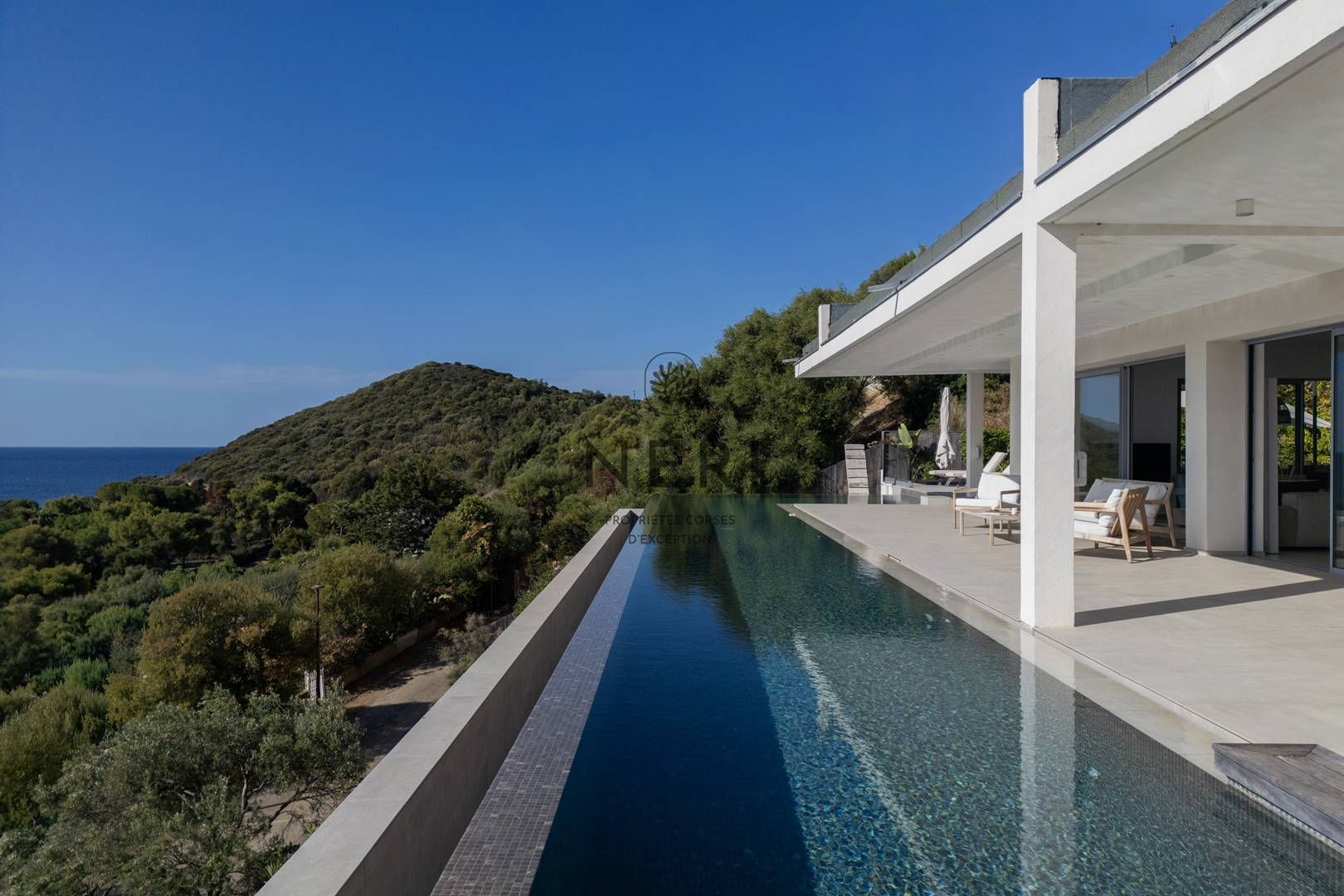 A modern villa with a long swimming pool, outdoor seating, and a view of the sea and hills.
