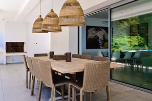 Un comedor moderno con una mesa de madera y sillas de mimbre, una parrilla empotrada y un ventanal con vistas al verdor.