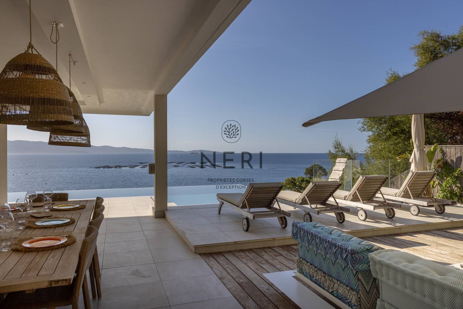 Un espacio de estar al aire libre techado con vista al mar, una mesa de comedor dispuesta con platos y vasos, y sillones de descanso.