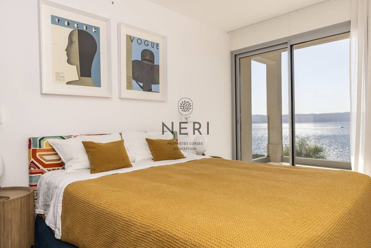 A bedroom with a large bed, two pillows, a colorful headboard, a framed poster on the wall, and a view of the sea through a sliding door.