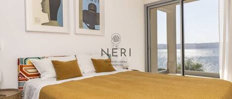 A bedroom with a large bed, two pillows, a colorful headboard, a framed poster on the wall, and a view of the sea through a sliding door.