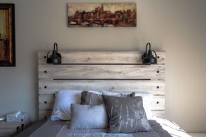 A bedroom with a wooden headboard, two bedside lamps, a bed with white and grey bedding, and a painting above the headboard. - Cute Condo with Pool View and lots of charm! (Houston)