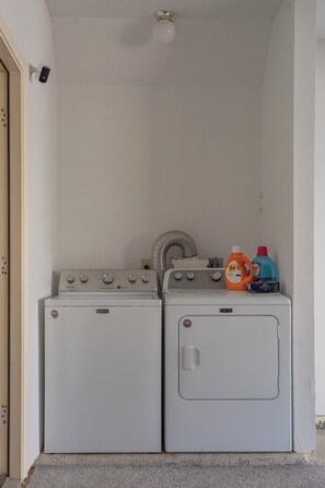 Dues sequedores Maytag blanques en una sala de bugaderia amb una paret blanca, una sortida de ventilació i amples amples de detergent per a la roba a la part superior.