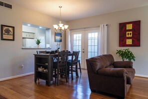 A modern living room with a brown leather sofa, a dining area with a dark wooden table and chairs, and a kitchen with white cabinets and a large window. - Cute Condo with Pool View and lots of charm! (Houston)