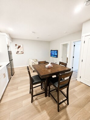 A modern kitchen with white cabinets, a wooden dining table, and a flat-screen TV mounted on the wall.