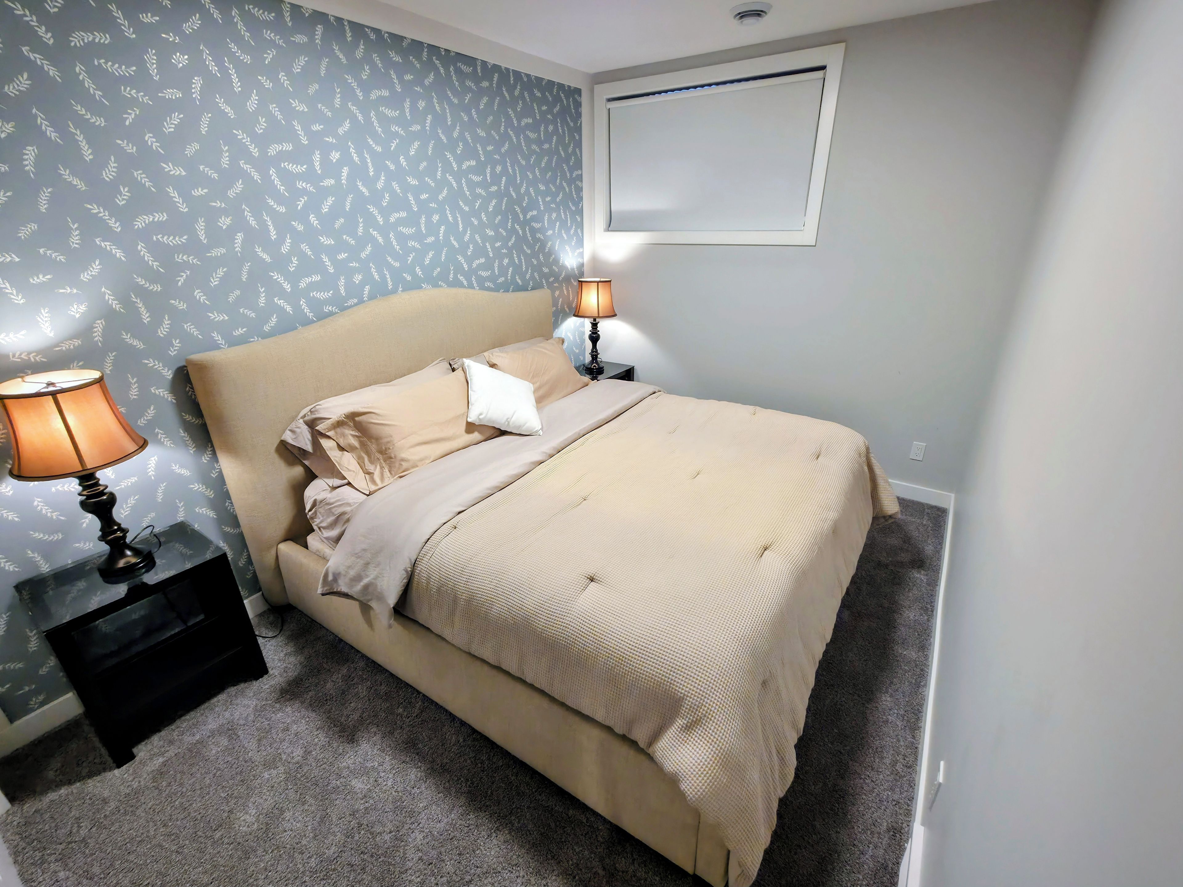 Quarto com cama, duas luminárias de cabeceira, mesa de cabeceira e papel de parede estampado.