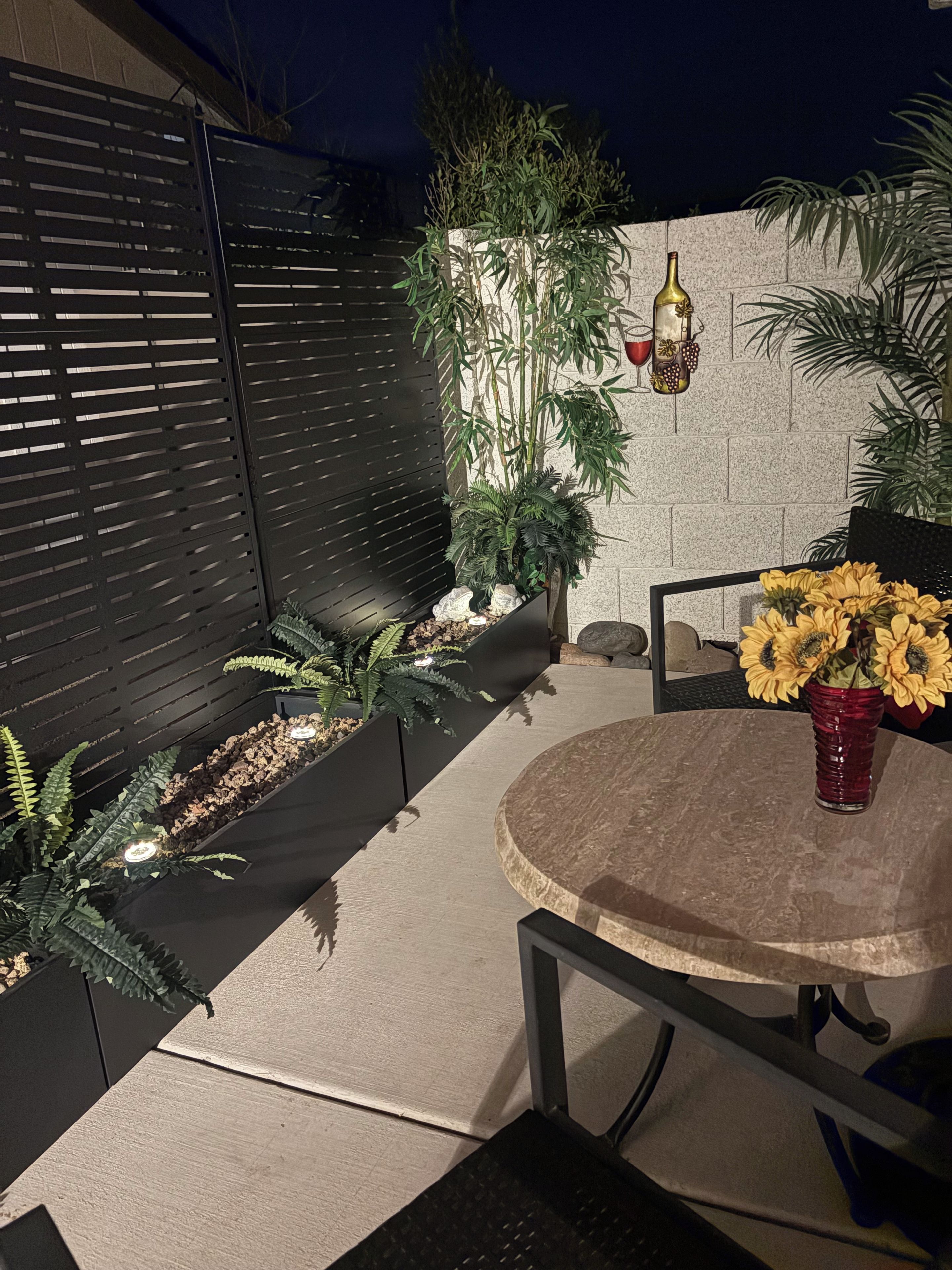 A patio area with a round table and chairs, surrounded by potted plants and a black fence.