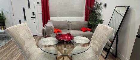 A modern dining area with a glass table, two chairs, a red decorative bowl, a grey sofa with red cushions, a potted plant, and a standing mirror.