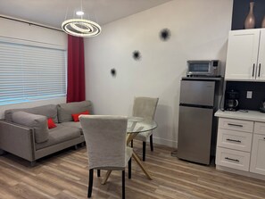 A modern living space with a grey sofa, a glass table, and a chair. A kitchen area with a refrigerator and microwave is visible. - Beautiful Guesthouse in Kierland. Studio A (Scottsdale)