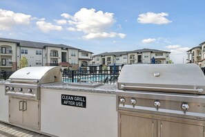 A clean grill area with a sign indicating it should be cleaned after use, a pool, and apartment buildings in the background.
