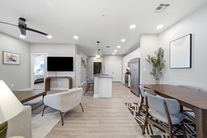 Eine moderne Küche mit einer zentralen Kochinsel, ein Essbereich mit Holztisch und -stühlen sowie ein Wohnbereich mit Sofa und Fernseher.