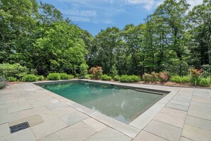 A rectangular swimming pool with a paved deck, surrounded by a black metal fence and lush greenery. - Upscale Fall Retreat | 3 Min URI + Outdoor Oasis (South Kingstown)