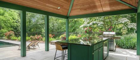 An outdoor patio with a wooden ceiling, green columns, a barbecue grill, and a pool.