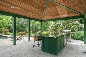 Um pátio externo com teto de madeira, colunas verdes, churrasqueira e piscina.