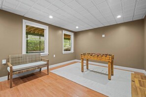 A room with a wooden foosball table, a wicker bench, and two windows with lattice screens. - Upscale Fall Retreat | 3 Min URI + Outdoor Oasis (South Kingstown)