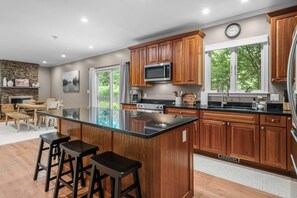 A modern kitchen with wooden cabinets, a large island with stools, a microwave, and a refrigerator. - Upscale Fall Retreat | 3 Min URI + Outdoor Oasis (South Kingstown)