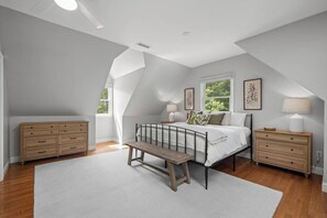 Ein Schlafzimmer mit schrägen Wänden, einem Bett mit Metallgestell, einer Holzbank, einer Kommode und einem Nachttisch mit Lampe.