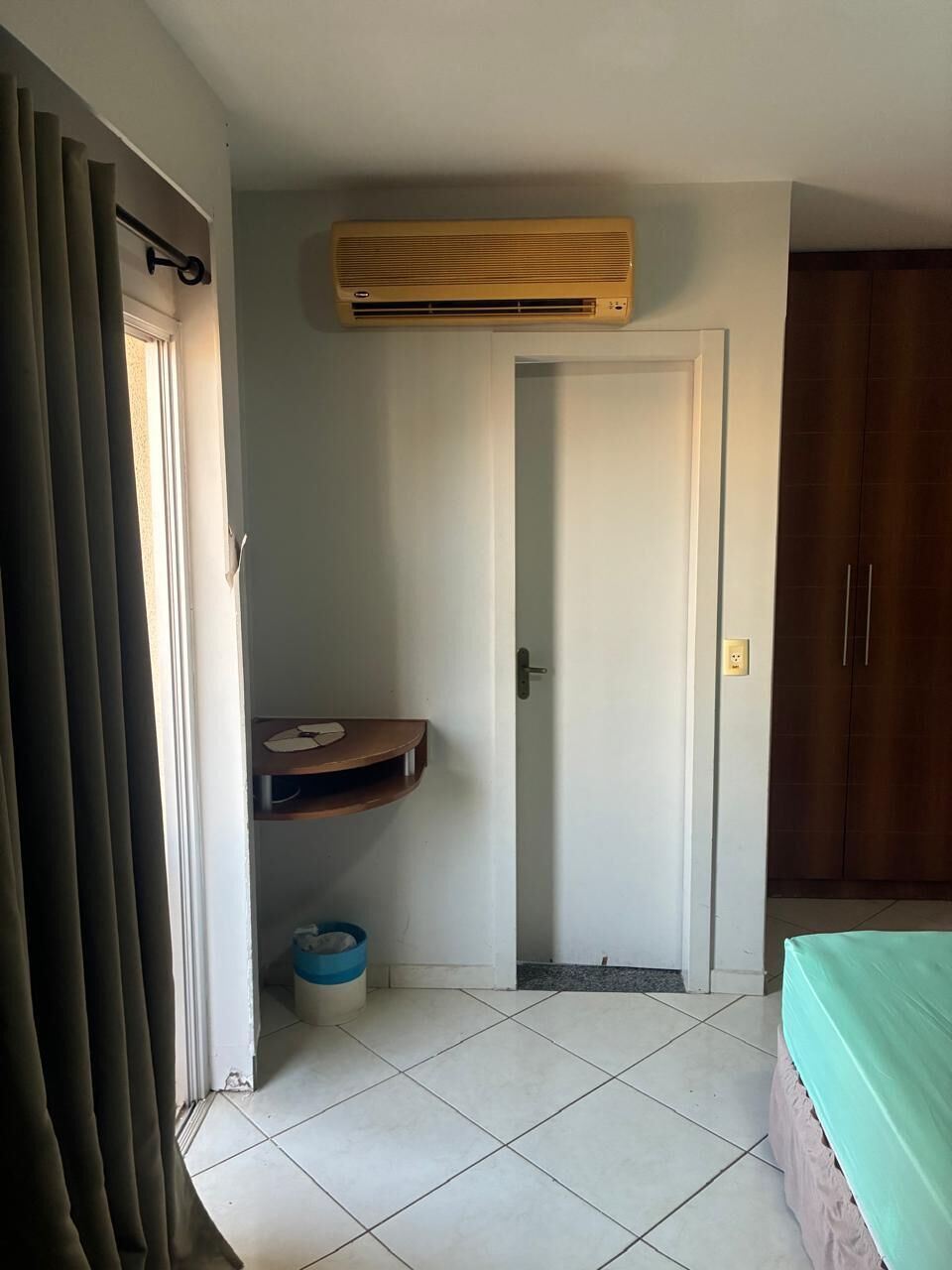 Een kamer met een bed, een deur, een airconditioningapparaat, een kleine prullenbak en een gordijn.