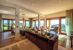 A spacious living room with a large sofa, multiple chairs, a coffee table, and large windows offering a view of the sea. - Tangaroa Estate – Luxury Coastal Escape (Waiheke Island)