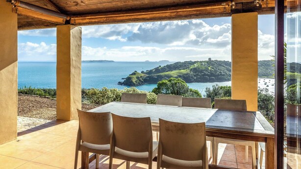 Ein überdachter Außenbereich mit Tisch und Stühlen mit Blick auf eine Küstenlandschaft mit Hügeln und dem Meer.
