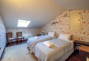 Ein Schlafzimmer mit zwei Betten, einem Oberlicht, einem Sessel, einem kleinen Tisch und floralem Wandpapier.