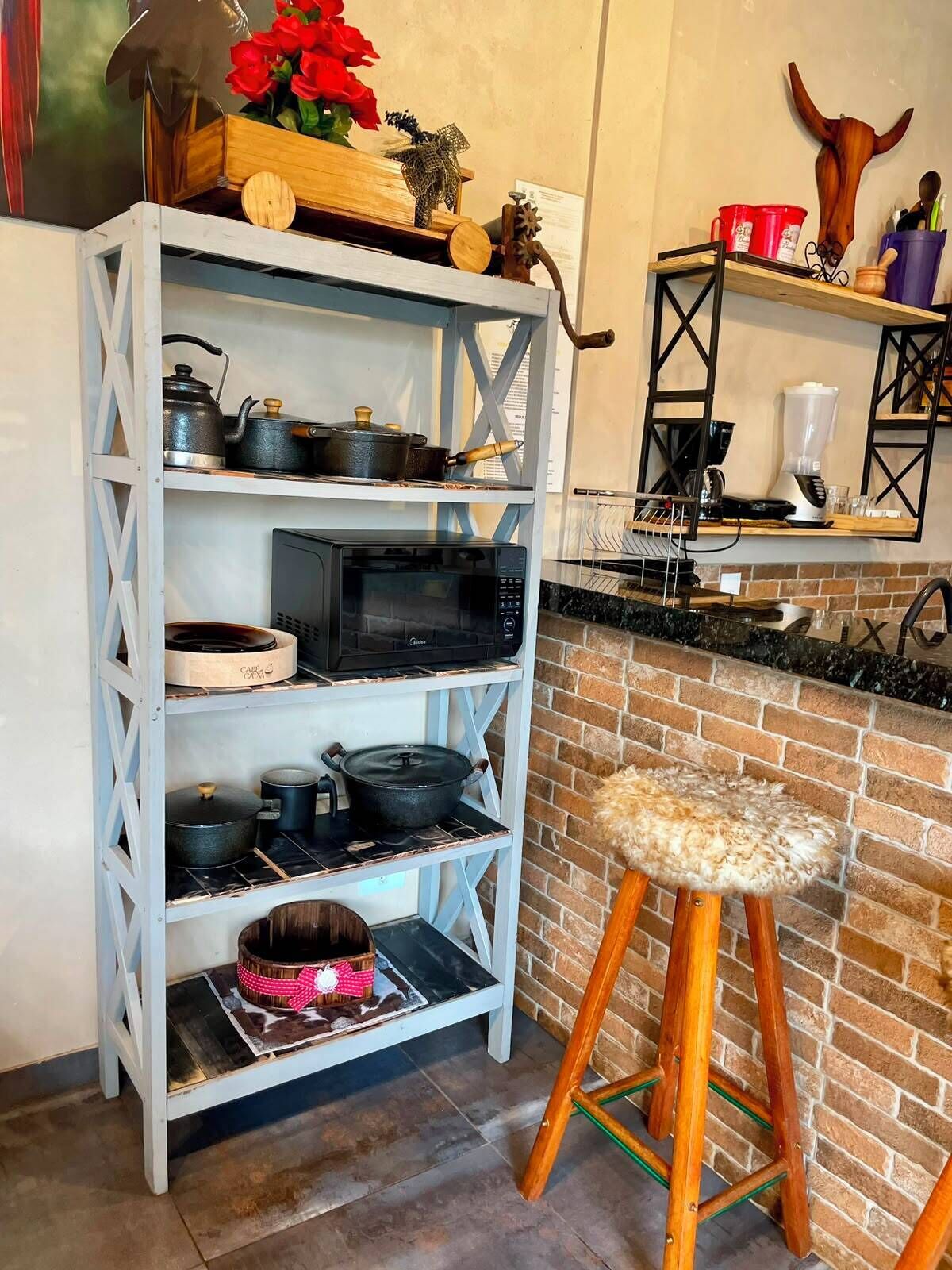 A kitchen with a white metal shelving unit holding pots and a microwave. There's a brick wall and a wooden stool with a fur seat.