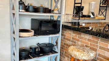 A kitchen with a white metal shelving unit holding pots and a microwave. There's a brick wall and a wooden stool with a fur seat.