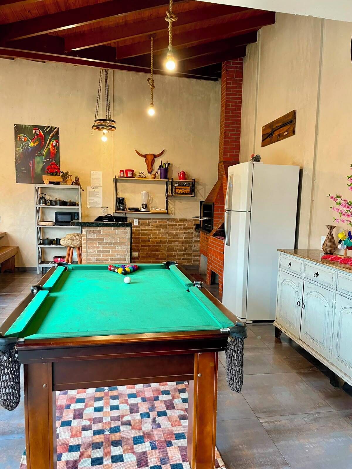 A pool table in a room with a brick wall, a refrigerator, and shelves with various items.