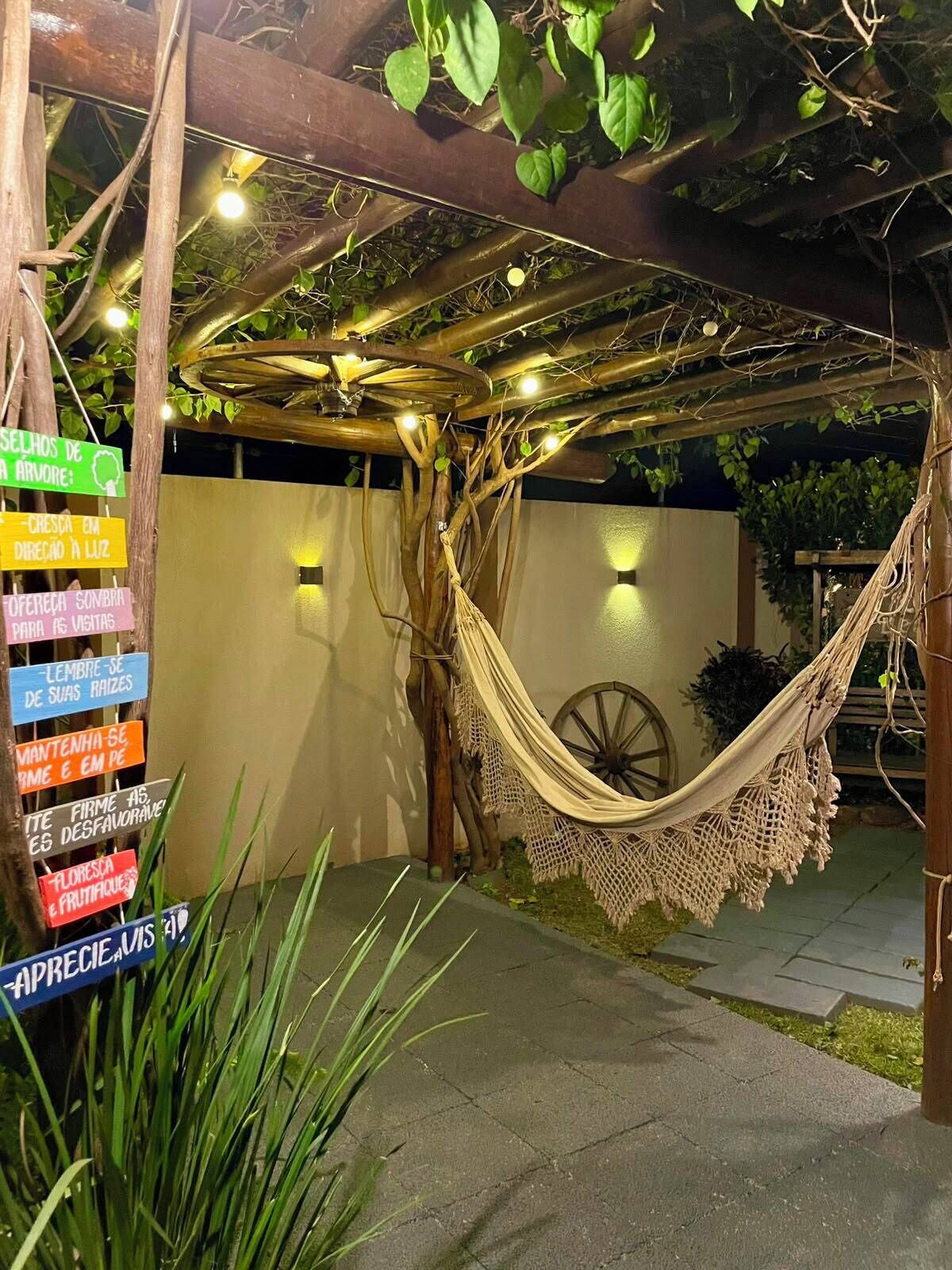 A hammock under a wooden pergola with string lights, surrounded by greenery and a rustic wheel.