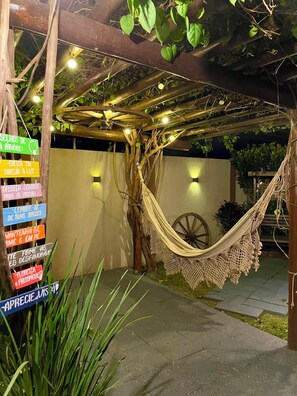 Uma rede sob uma pérgola de madeira com luzes de corda, rodeada por vegetação e um carro de roda rústico.