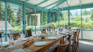 A conservatory with a dining table set for a meal, surrounded by large windows offering a view of greenery and a mountain.