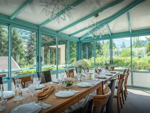 A conservatory with a dining table set for a meal, surrounded by large windows offering a view of greenery and a mountain.