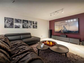A modern living room with a sectional sofa, a flat-screen TV mounted on the wall, and a coffee table with a tray of orange juice.