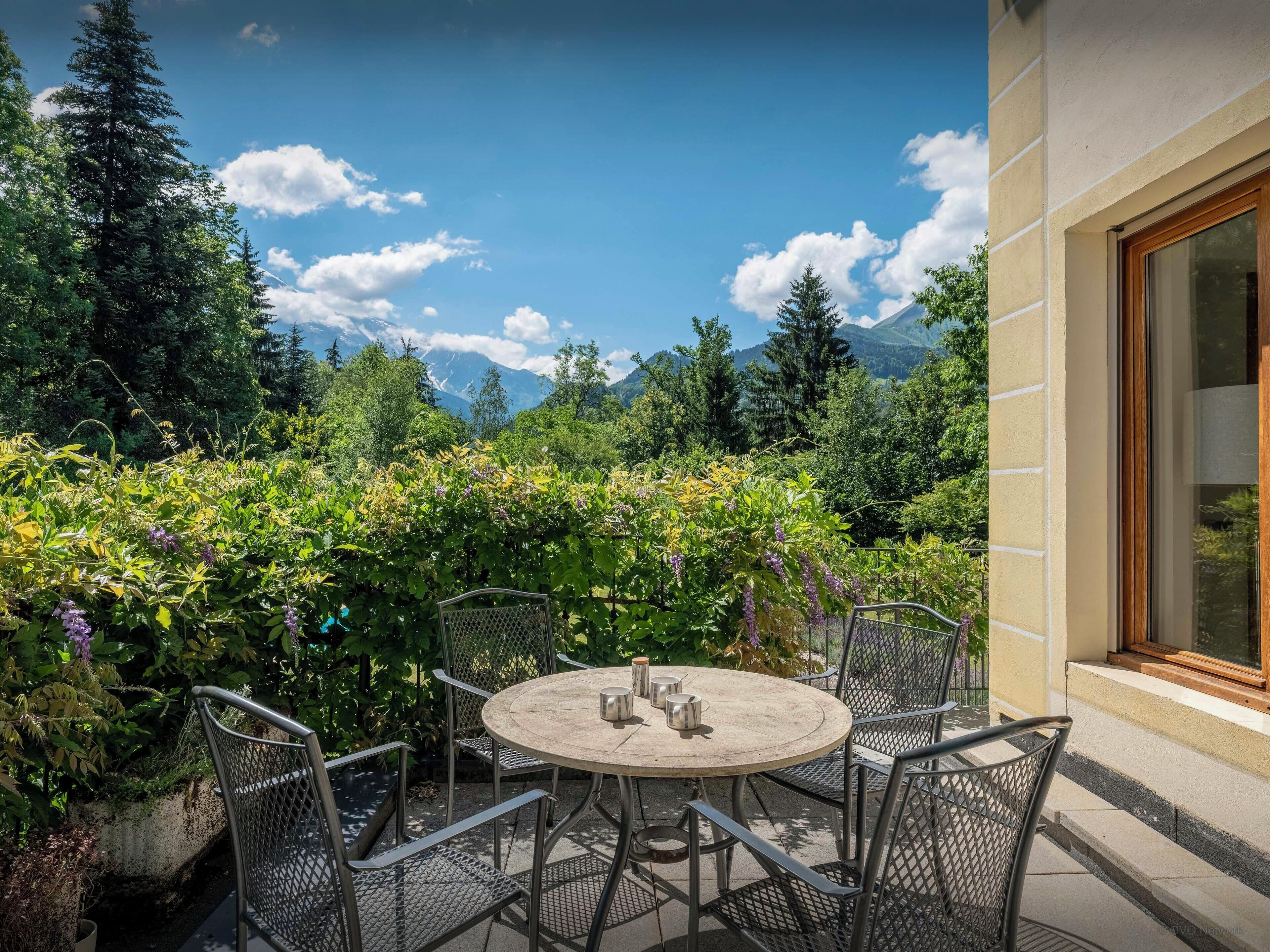 Eine Terrasse im Freien mit einem runden Tisch und Metallstühlen, umgeben von üppiger Bepflanzung und Blick auf Berge.