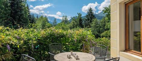 Eine Terrasse im Freien mit einem runden Tisch und Metallstühlen, umgeben von üppiger Bepflanzung und Blick auf Berge.