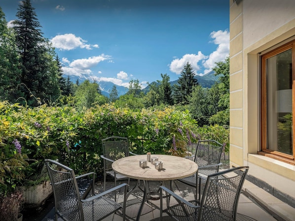 An outdoor patio with a round table and metal chairs, surrounded by lush greenery and a mountain view.