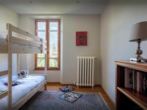 A room with a bunk bed, a radiator, a bookshelf, and a window with a view of greenery.