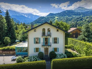 Ein zweigeschossiges Haus mit Balkon, umgeben von üppigem Grün und Bergen im Hintergrund.