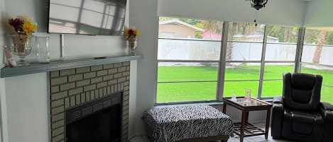 Una sala de estar con chimenea, un televisor montado sobre ella, un ventilador de techo y un ventanal que ofrece vista a un patio trasero.