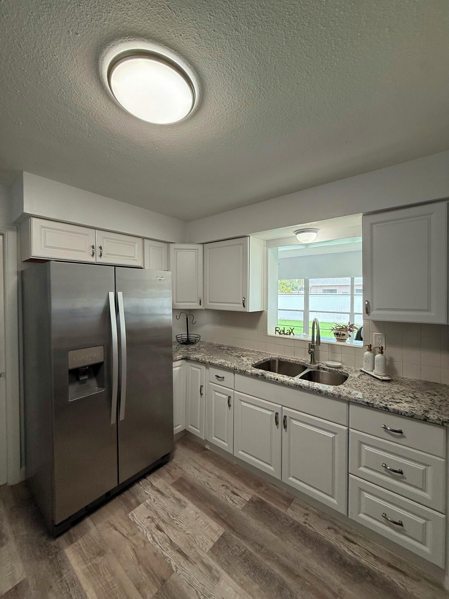 A kitchen with white cabinets, a stainless steel refrigerator, a granite countertop, and a window with a view.